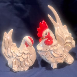 May include: A pair of white ceramic rooster and hen figurines with gold accents. The rooster has a red comb and wattle. The hen has a red comb.