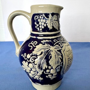 May include: A stoneware pitcher with a dark blue body and a light beige rim, handle, and base. The pitcher features embossed grape and leaf designs. The handle is curved, and the spout is small. The pitcher has a capacity of approximately 1 liter.