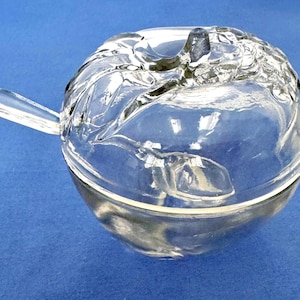 Może przedstawiać: Przezroczysta szklana miska w kształcie jabłka z pokrywką i uchwytem. Pokrywka ma teksturowany wzór, a uchwyt to małe, zakrzywione przedłużenie. Miska jest ustawiona na niebieskim tle.