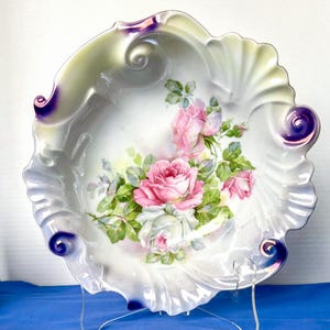 May include: A white porcelain bowl with a scalloped edge and a floral design featuring pink and white roses. The bowl has a purple iridescent glaze on the edges.