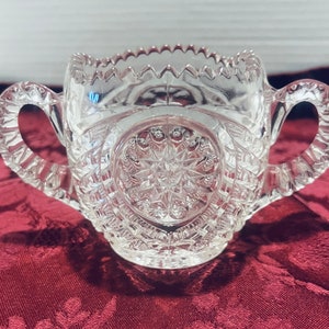 May include: A clear glass sugar bowl with two handles. The bowl has a cut glass design with a starburst pattern in the center.