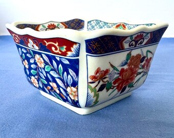 Peut inclure: Bol en céramique carré avec un bord festonné. Le bol présente une base blanche avec des motifs floraux et de feuilles bleus, rouges et dorés. Les coins sont légèrement arrondis, et le bol repose sur une surface bleue.
