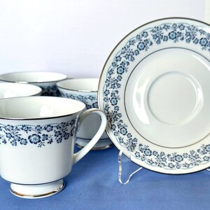 Può includere: Servizio di tazze e piattini bianchi con motivo floreale blu. Il set comprende quattro tazze e un piattino. Le tazze hanno un bordo dorato sulla base e sul bordo. Il piattino ha un motivo floreale abbinato e un bordo dorato.