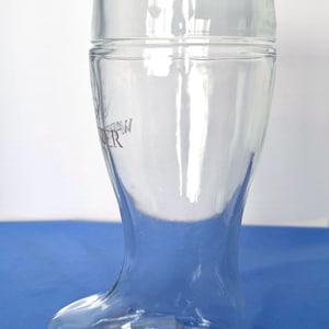 May include: A clear glass beer boot with a rounded top and a foot-shaped base. The glass has a curved design and a slight taper. The text "AW ER" is visible on the side. The boot-shaped glass is designed for beer.