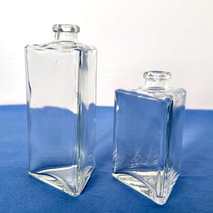 Könnte beinhalten: Zwei klare Glasflaschen mit einem einzigartigen, kantigen Design. Die Flaschen haben eine rechteckige Form mit dreieckigem Boden und kleinem, abgerundetem Hals. Die Flaschen sind leer und scheinen aus dickem Glas zu sein. Der Hintergrund ist einfarbig blau.