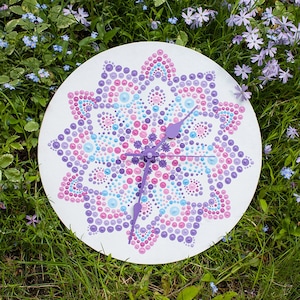 May include: A white clock with a purple, pink, and blue mandala design. The clock is surrounded by green grass and purple flowers.