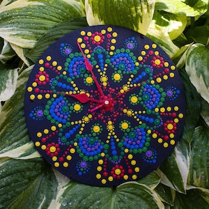 May include: A black circular clock with a colorful mandala design painted on it. The clock is surrounded by green leaves with white edges.