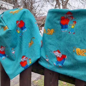 Könnte beinhalten: Türkisfarbenes Mützen- und Halstuch-Set mit einem verspielten Muster aus Elefanten, Tigern, Löwen und einem Kind. Der Elefant trägt ein rotes Hemd und blaue Hosen. Die Mütze ist umgeschlagen.