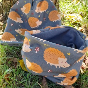 Könnte beinhalten: Ein Set aus Mütze und Halstuch in Blau. Beide Teile sind mit einem sich wiederholenden Muster von Cartoon-Igeln in Braun- und Beigetönen versehen. Das Design umfasst kleine Blumen- und Herzmotive.