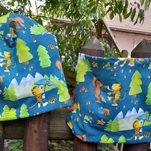 Könnte beinhalten: Zwei blaue Stoffmützen mit einem Waldtier- und Waldmuster. Eine Mütze ist eine Beanie und die andere ein Halstuch. Beide Mützen zeigen einen Tiger, einen Bären und Bäume.