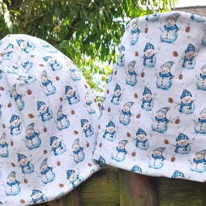 Könnte beinhalten: Zwei weiße Mützen mit einem wiederkehrenden Aquarellmuster aus Schneemännern, die blau gestreifte Mützen und Schals tragen, durchsetzt mit kleinen braunen Tannenzapfen. Der Stoff hat eine weiche Textur und ein durchgehendes Muster auf beiden Artikeln.