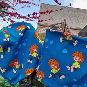 Könnte beinhalten: Zwei blaue Stoffartikel mit einem sich wiederholenden Muster eines rothaarigen Comic-Charakters, der ein gelbes Hemd und eine grüne Hose trägt. Der Charakter rennt und hält ein gelbes Objekt in der Hand. Die Stoffartikel hängen an einem Holzzaun.