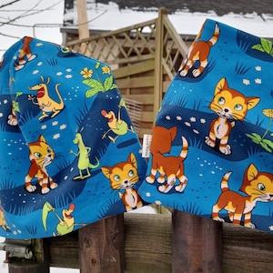 Könnte beinhalten: Zwei blaue Stoffartikel mit einem sich wiederholenden Muster aus Cartoon-Katzen, Kängurus und anderen Kreaturen in einer grasbewachsenen Landschaft. Der Hintergrund ist dunkelblau mit weißen Blumenakzenten. Bei den Artikeln handelt es sich wahrscheinlich um eine Mütze und einen Schal.