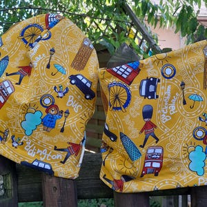 Könnte beinhalten: Zwei gelbe Stoffartikel mit London-Motiv. Das Design umfasst Illustrationen von London Eye, Big Ben, roten Bussen und einem Bären in einem blauen Mantel. Text enthält "Paddington" und "Bus".