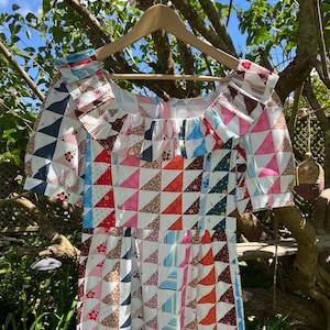 Puede incluir: Un vestido blanco con un diseño de patchwork con una variedad de colores, incluyendo rojo, azul, rosa y marrón. El vestido tiene un escote cuadrado y mangas cortas. El vestido está colgado en una percha de madera.