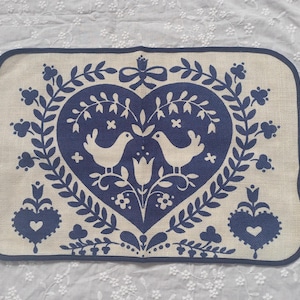 Puede incluir: Mantel individual rectangular beige con un diseño en forma de corazón azul marino con dos pájaros, elementos florales y un borde. El diseño es simétrico y evoca una estética de arte popular.