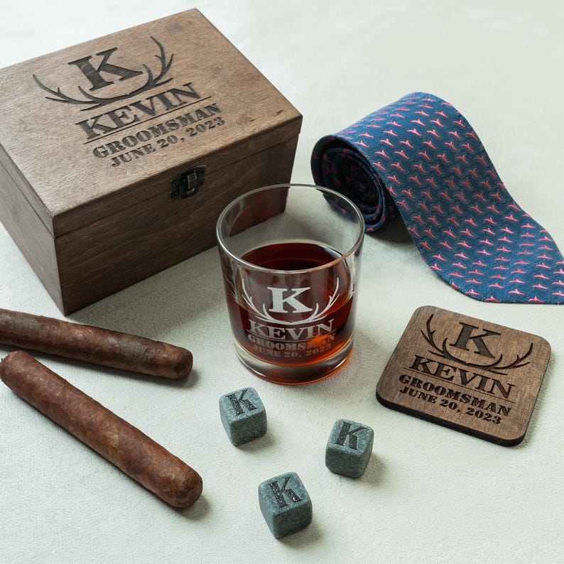 May include: A personalised groomsman gift set including a wooden box, a rocks glass with whiskey, a tie, a coaster, and whiskey stones. The box, glass, and coaster are engraved with the name "KEVIN" and the date "JUNE 20, 2023". Two cigars are also included.