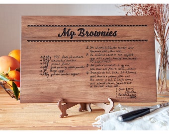 Könnte beinhalten: Ein Holzbrett zum Schneiden mit einem handgeschriebenen Rezept für Brownies, das darauf graviert ist. Das Rezept enthält Zutaten wie dunkle Schokolade, ungesalzene Butter, Mehl, Kakaopulver, Eier und braunen Zucker. Das Brett hat einen dekorativen Rand und den Text "My Brownies" oben.