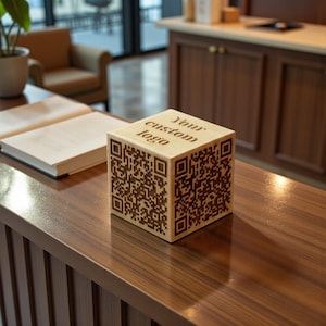 Könnte beinhalten: Ein helles Holzwürfel mit QR-Codes auf vier Seiten und dem Text "Your custom logo" auf der Oberseite eingraviert. Der Würfel steht auf einer dunklen Holzoberfläche. Werbeartikel.