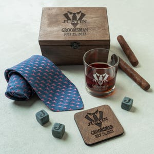 May include: A wooden box, a blue tie with pink accents, a rocks glass with whiskey, cigars, and stone cubes. The box, glass, and coaster are engraved with "JUSTIN GROOMSMAN JULY 25, 2023".