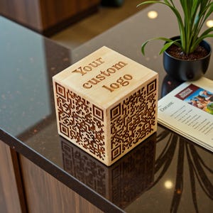 May include: A light-colored wooden cube featuring a laser-engraved QR code on its side faces and the text "Your custom logo" engraved on the top surface. The cube has a smooth, natural wood grain finish and sharp, clean edges on all sides.