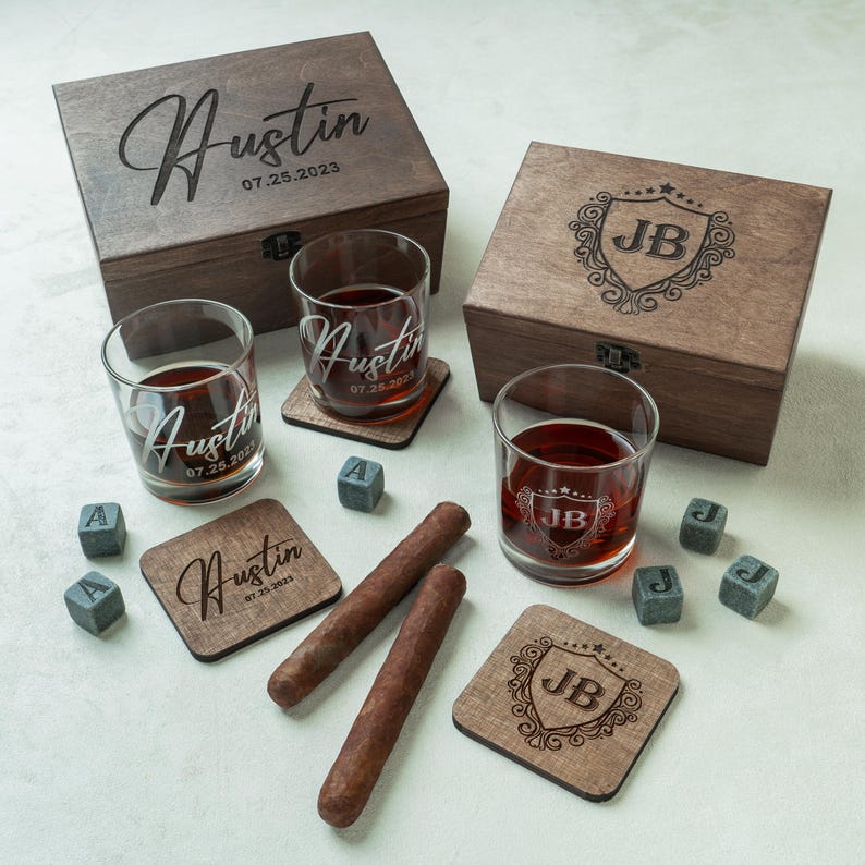 May include: A wooden gift set featuring engraved boxes, whiskey glasses, coasters, and whiskey stones. The glasses and coasters are personalized with names and dates. Two cigars are also included.