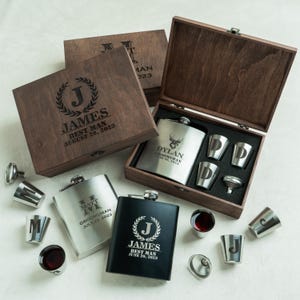May include: A collection of personalized flask sets in wooden boxes. The sets include stainless steel flasks, shot glasses, and funnels. The boxes and flasks are engraved with names, initials, and dates. The flasks are black and silver.