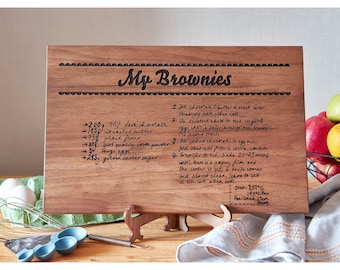 Könnte beinhalten: Ein Holzbrett zum Schneiden mit einem handgeschriebenen Rezept für Brownies. Das Rezept enthält Zutaten und Anweisungen zum Backen von Brownies. Das Brett hat einen dekorativen Rand und steht auf einem Ständer.