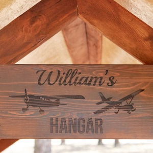 May include: A wooden sign with the text "William's Hangar" in a dark brown font. The sign is decorated with two small airplane silhouettes and four propellers.