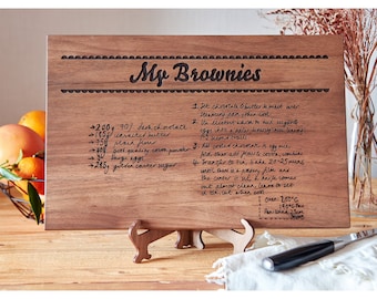 Könnte beinhalten: Ein Holzbrett zum Schneiden mit einem handgeschriebenen Rezept für Brownies, das darauf graviert ist. Das Rezept enthält Zutaten wie dunkle Schokolade, ungesalzene Butter, Mehl, Kakaopulver, Eier und braunen Zucker. Das Brett hat einen dekorativen Rand und den Text "My Brownies" oben.