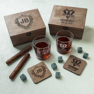 May include: A wooden gift set featuring two glasses of amber liquid, two cigars, and engraved wooden coasters and boxes. The boxes and coasters are engraved with initials and the text "Groomsman" and a date. Grey stone cubes are included.