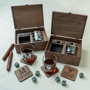 May include: Two wooden boxes containing engraved whiskey glasses, coasters, and ice cubes. The glasses and coasters feature personalized text and a deer head design. The boxes are open, revealing the contents, and cigars are placed on the side.