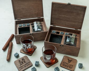 May include: Two wooden boxes with engraved whiskey glasses, coasters, and ice cubes. The glasses and coasters feature personalized text and a deer head design. The boxes are open, revealing the contents, and cigars are placed on the side.