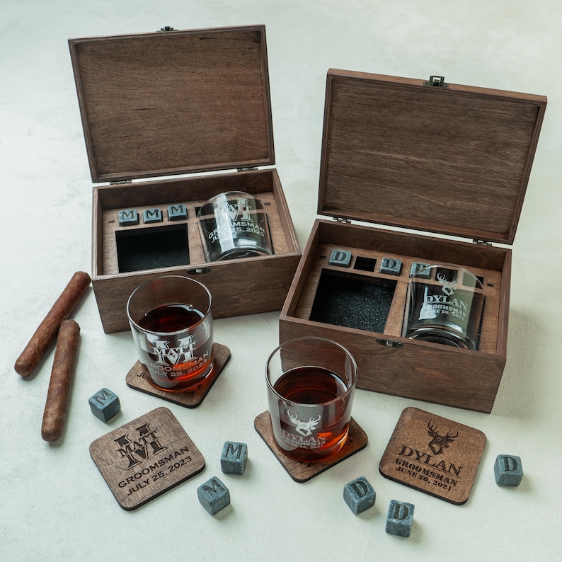 May include: Two wooden boxes containing engraved whiskey glasses, coasters, and ice cubes. The glasses and coasters feature personalized text and a deer head design. The boxes are open, revealing the contents, and cigars are placed on the side.