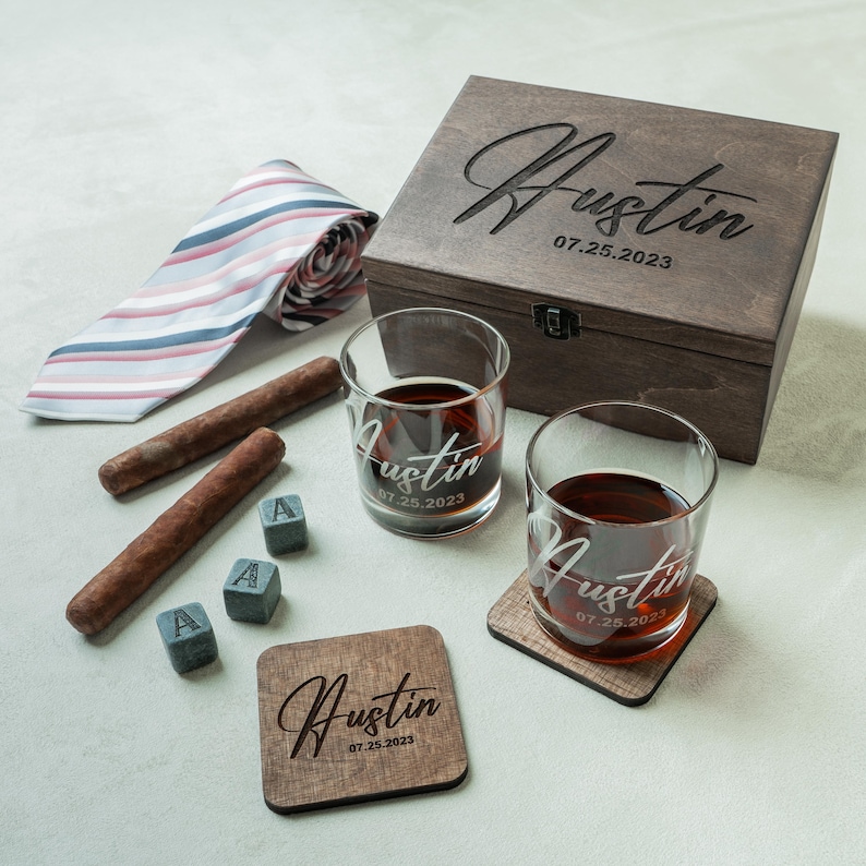 May include: A personalised gift set with a wooden box, two whiskey glasses, coasters, and ice cubes. The set includes a striped tie and cigars. The box, glasses, and coasters are engraved with the name "Austin" and the date "07.25.2023".