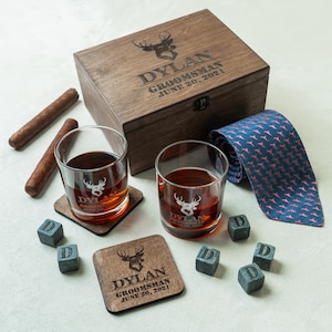 May include: A wooden gift box with the text "DYLAN GROOMSMAN JUNE 20, 2021" and a deer head design. Includes two whiskey glasses, coasters, ice cubes, a blue tie with a pink pattern, and two cigars.