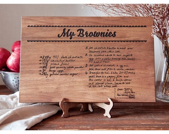 Könnte beinhalten: Ein Holzbrett zum Schneiden mit einem handgeschriebenen Rezept für Brownies. Das Rezept enthält Zutaten wie dunkle Schokolade, ungesalzene Butter, Mehl, Kakaopulver, Eier und braunen Zucker. Das Brett hat einen dekorativen Rand und der Text "My Brownies" ist oben eingraviert.