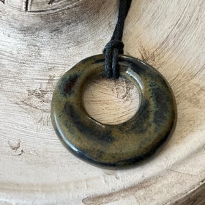 May include: A ceramic pendant necklace featuring a circular, donut-shaped pendant. The pendant has a glossy finish with green, brown, and blue hues. It is suspended from a black cord. The necklace is displayed on a textured, off-white surface.