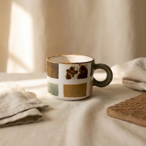 May include: A ceramic mug featuring a white base with a grid pattern of rectangular patches in shades of brown, rust, and muted green. The mug has a thick, rounded handle and a smooth, cylindrical body with a speckled, textured finish.