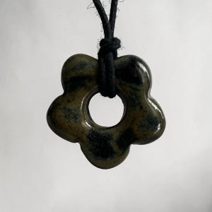 Könnte beinhalten: Ein Keramikanhänger in Form einer fünfblättrigen Blume mit einer glänzenden, gesprenkelten dunkelgrünen und schwarzen Glasur. Der Anhänger hat einen kreisförmigen Ausschnitt in der Mitte und hängt an einer einfachen schwarzen Kordel.