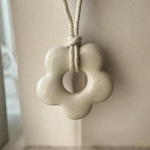 May include: A smooth, cream-colored ceramic pendant shaped like a five-petaled flower with a central circular cutout. The pendant is suspended from a thick, braided beige cord that is knotted securely at the top of the ceramic flower shape.