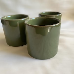 May include: Three small, cylindrical ceramic cups with a smooth, glossy olive green glaze. Each cup features a simple, handleless design with a uniform finish and a slightly rounded rim, arranged in a cluster to show their matching appearance.