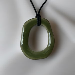Könnte beinhalten: Eine Anhängerkette mit einem unregelmäßig geformten, organischen Ring aus olivgrünem Glas, der an einer einfachen schwarzen Kordel hängt. Die glatte, glänzende Oberfläche des grünen Glases reflektiert das Licht und bildet einen Kontrast zur dunklen, matten Kordel.
