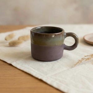 May include: A ceramic mug featuring a two-tone design with a matte dark purple base and a glossy, smooth olive green upper section. The mug has a wide, rounded handle on the right side and a straight, cylindrical body with a flat bottom edge.