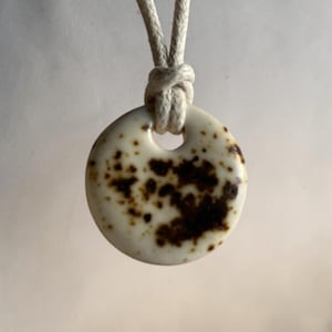 May include: A circular pendant with a smooth, off-white surface featuring irregular dark brown speckles and patches. The pendant has a hole at the top through which a thick, light-colored woven cord is looped and knotted to form a necklace.