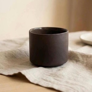 May include: A dark brown, cylindrical ceramic cup with a matte finish. The vessel features straight, vertical sides and a flat base, presenting a simple, unadorned design with a consistent, deep earthy tone throughout the exterior surface.