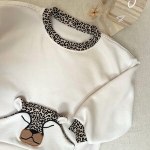 Peut inclure: Sweat-shirt blanc avec un col, des poignets et un motif de visage de léopard. Le sweat-shirt a une coupe ample, probablement pour un enfant. Le motif comprend un nez beige et des yeux fermés.