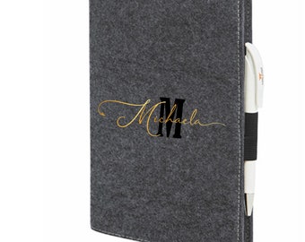May include: Gray felt notebook with a black and gold monogram of the letter "M" and the name "Michaela" in gold script. The notebook has a pen holder on the side.