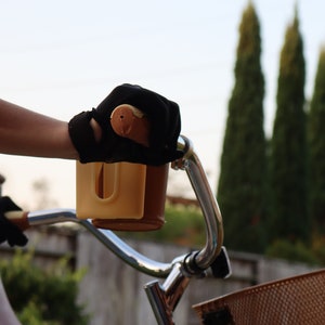 May include: A person riding a bicycle with a brown basket and a beige handlebar cup holder. The person is wearing black gloves.