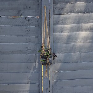 Puede incluir: Un macetero colgante de macramé con una maceta de cerámica gris oscuro llena de plantas verdes y moradas. La maceta tiene un diseño geométrico blanco y negro. El macetero está suspendido de una valla de madera gris.